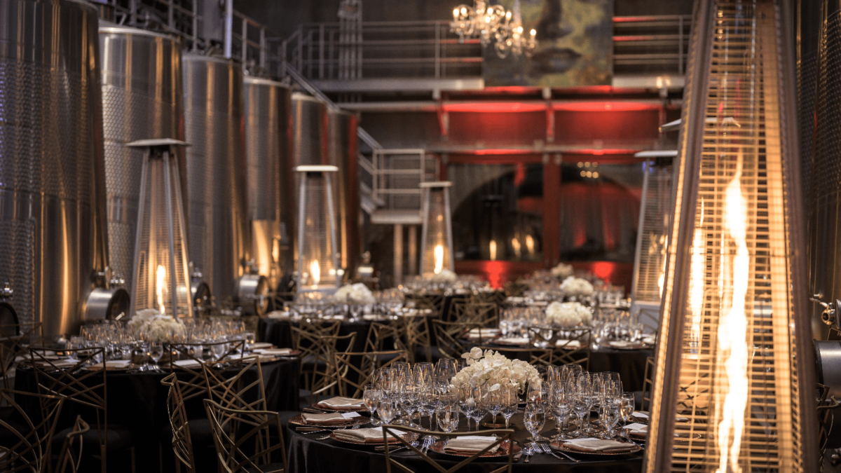 A sophisticated setup for one of the best annual events in Napa Valley, featuring an industrial-style venue with large metal tanks and red lighting. Elegant tables boast white floral arrangements, neatly arranged silverware, glassware, and black tablecloths. Tall heaters and chandeliers enhance the ambiance.