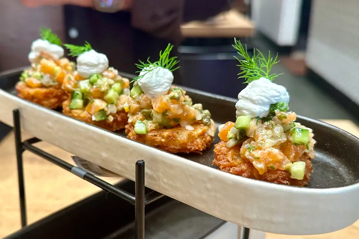 A rectangular dish holds four bite-sized appetizers. Each appetizer consists of a crispy base topped with finely diced vegetables, a dollop of cream, and garnished with a sprig of fresh dill. Perfect for those wondering where to eat Bay Area delicacies, the dish is presented on a sleek metal stand.