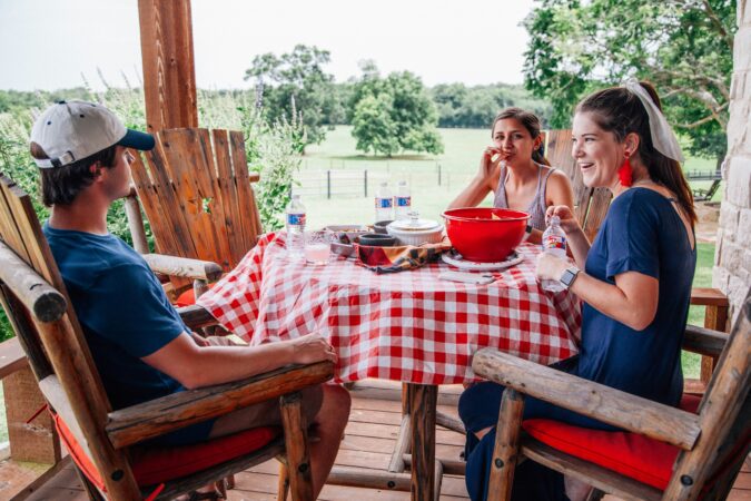 Three people are sitting around a table on a porch, enjoying a meal. The table is covered with a red and white checkered tablecloth and has various dishes reminiscent of Fourth of July celebrations. The background features a green field and trees. Everyone is casually dressed and smiling.