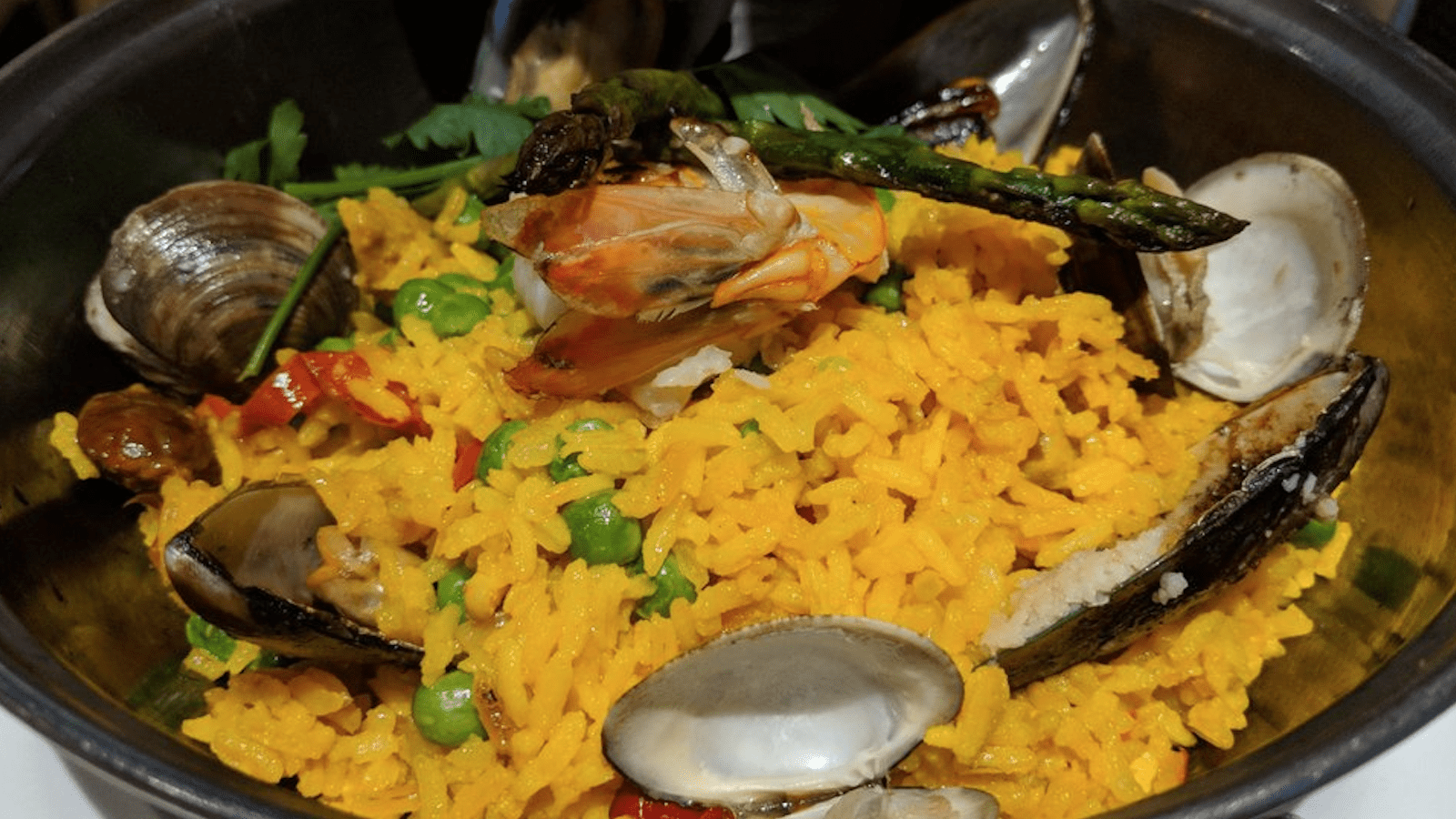 A vibrant dish of paella featuring bright yellow rice mixed with peas and red peppers. The dish, perfect for the best lunch on the Monterey Peninsula, is topped with clams, mussels, a shrimp, and garnished with a green herb and green beans.