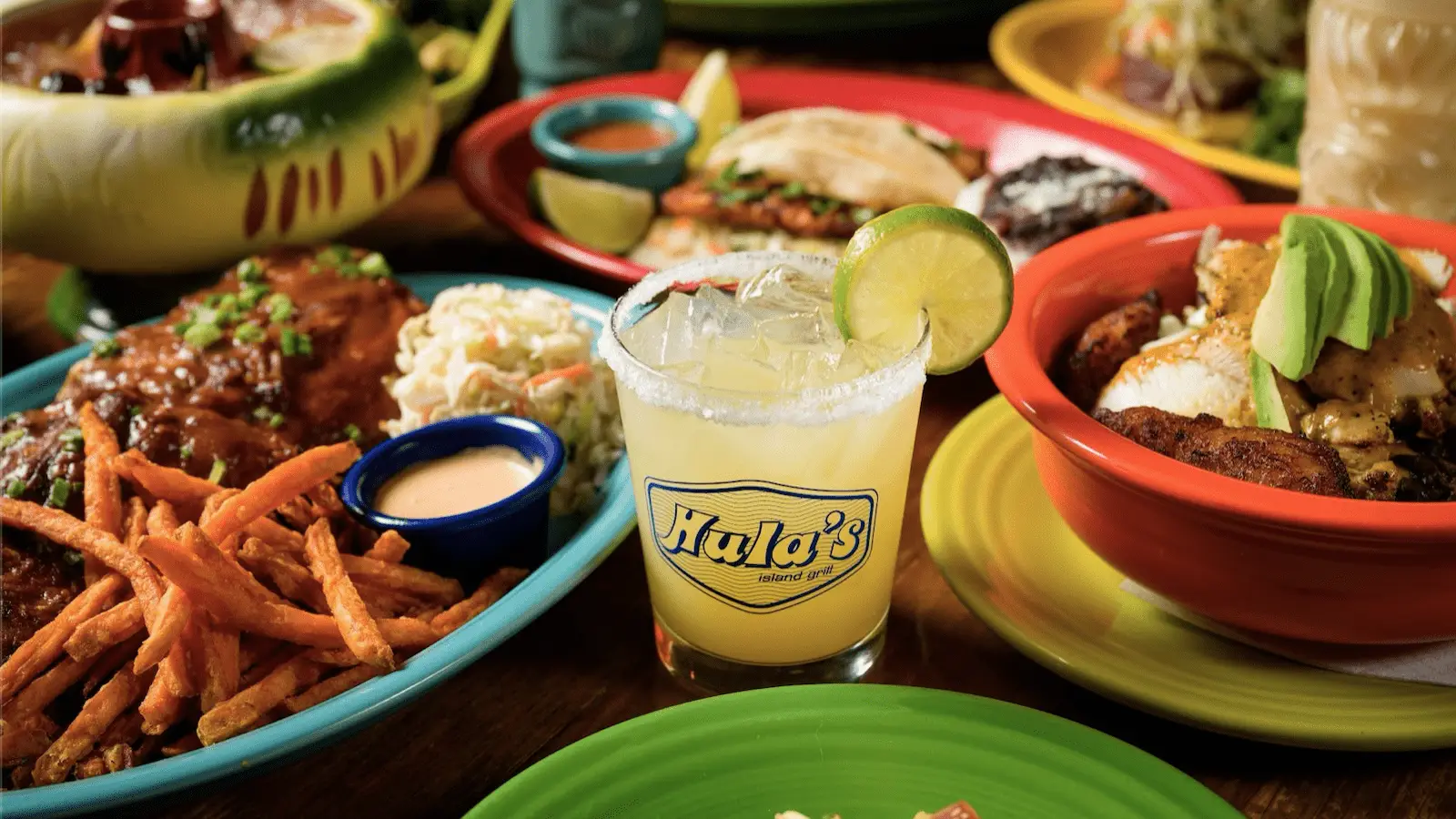 A table filled with various dishes, including sweet potato fries with dip, a bowl of rice topped with avocado and other vegetables, another plate with coleslaw and ribs, and a glass of a yellow drink garnished with a lime slice. For the best lunch in the Monterey Peninsula, look for the "Hula's" logo on the glass.