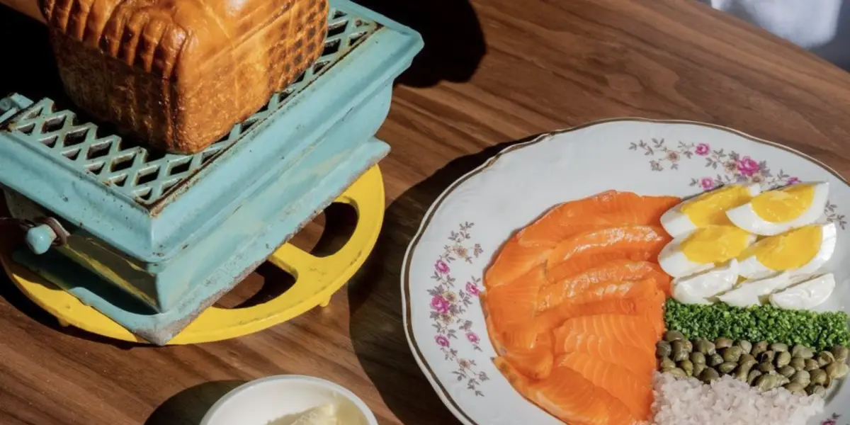 A dish on a wooden table with thinly sliced salmon, quartered boiled eggs, capers, and a small portion of rice, served on a floral-patterned plate. Next to the dish is a loaf of bread on an ornate, vintage-style green and yellow grill; truly the best lunch on the Monterey Peninsula.