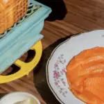 A dish on a wooden table with thinly sliced salmon, quartered boiled eggs, capers, and a small portion of rice, served on a floral-patterned plate. Next to the dish is a loaf of bread on an ornate, vintage-style green and yellow grill; truly the best lunch on the Monterey Peninsula.