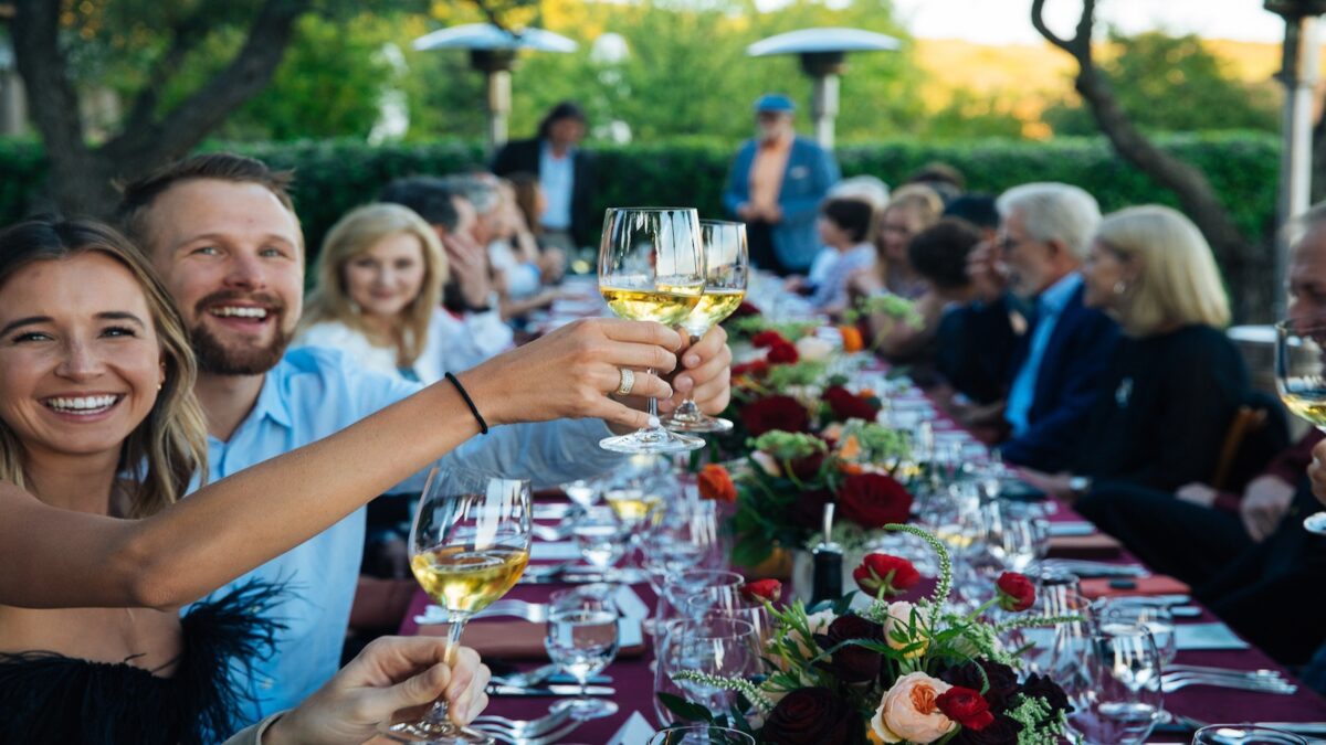 A group of people seated at an elegantly set outdoor table in Napa Valley, raising their wine glasses in a toast. The table is adorned with red tablecloths, floral arrangements, and set with glassware and plates. The background features greenery and patio heaters. Everyone looks cheerful at one of the best annual events.