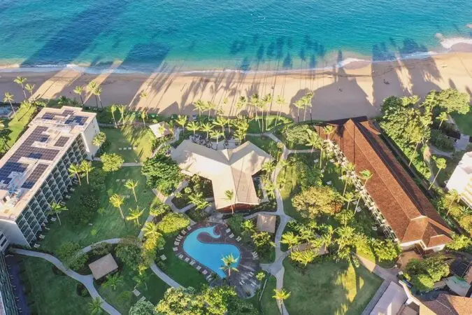 Aerial view of a beachfront resort in Ka‘anapali featuring lush greenery, a pool shaped like a fish, and multiple buildings including a large traditional-style structure with a slanted roof. The beach and turquoise ocean are visible in the background, with shadows of tall palm trees cast on the sand. Visit Ka‘anapali!