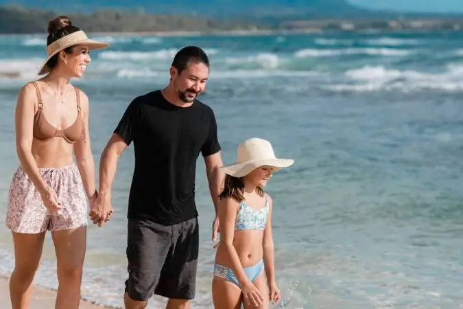 A woman, a man, and a girl walk along a sandy beach by the ocean. The woman is wearing a bikini top, shorts, and a visor; the man is in a black t-shirt and shorts; and the girl is in a two-piece swimsuit and sun hat. They are smiling and holding hands, enjoying their time at Outrigger Kauai Beach Resort & Spa.