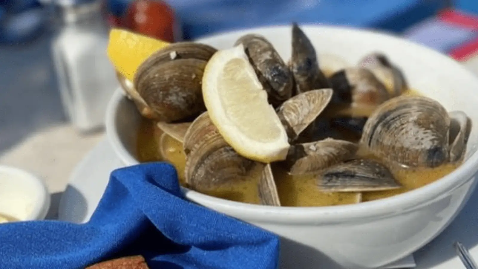 A white bowl filled with steamed clams in a broth is garnished with lemon wedges. Resting on a blue napkin with a salt shaker blurred in the background, it epitomizes the best lunch experience on the Monterey Peninsula, enhanced by an inviting outdoor setting.