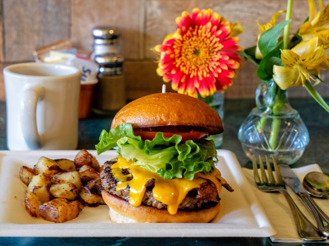 A cheeseburger with lettuce and tomato on a bun is served on a white plate with a side of roasted potatoes, showcasing why it’s among the Best Burgers in Mendocino. A white mug, salt and pepper shakers, and two vases with vibrant flowers are in the background on a green table. Fork, knife, and spoon are placed on a napkin.