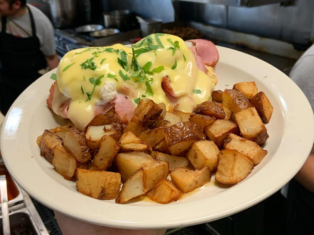 A plate of eggs Benedict, touted as the best breakfast in Mendocino, features poached eggs topped with hollandaise sauce and garnished with chopped parsley, resting on slices of ham over English muffins. Golden-brown, cubed breakfast potatoes are served alongside as a side dish.