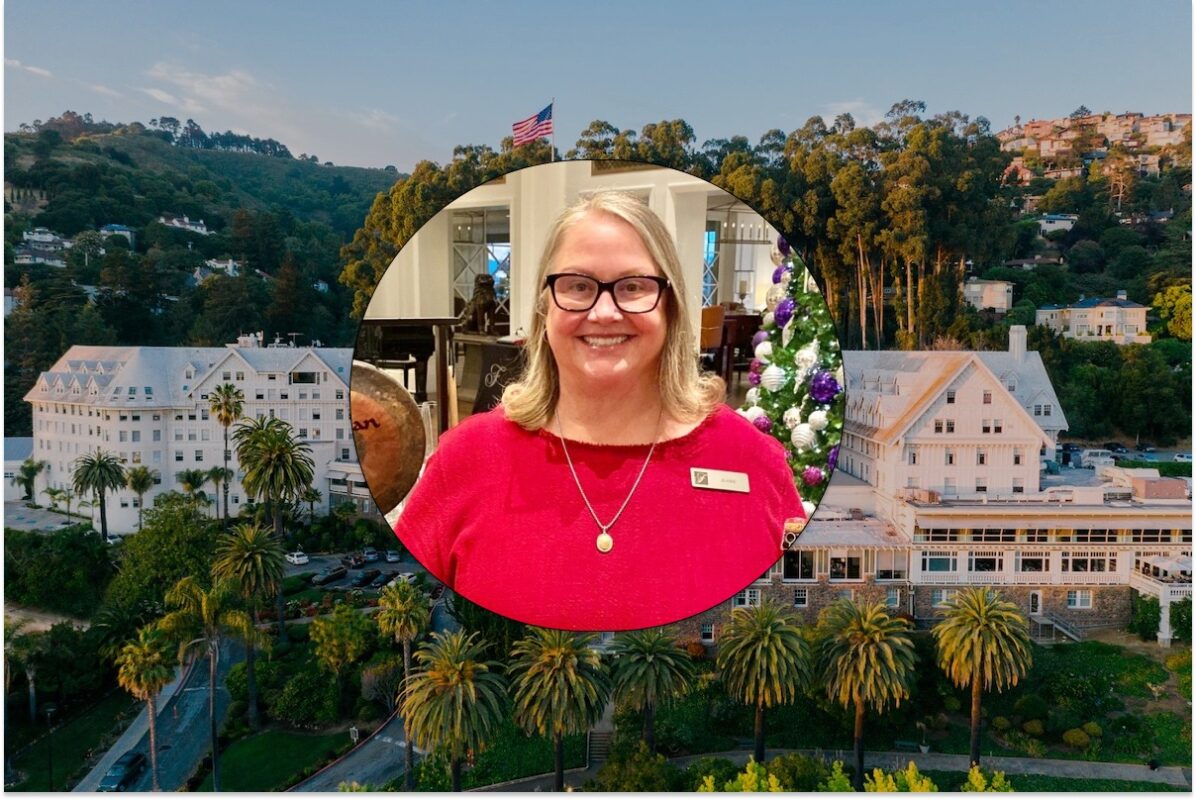 A woman with blonde hair and glasses smiles at the camera, wearing a red shirt and a nametag. Behind her, an outdoor scene captured through a circular frame shows a resort-like area with buildings, lush greenery, and an American flag flying on a hill—an inviting snapshot urging all to visit East Bay.