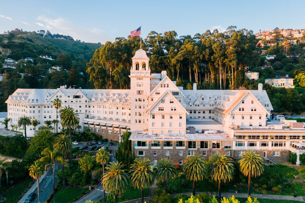 A large, historic white building with a central tower, displaying an American flag at the top, set against a backdrop of lush green hills and tall trees. Palm trees and neatly manicured gardens surround the property. Houses are visible on the hillside. Visit East Bay to explore its beauty!