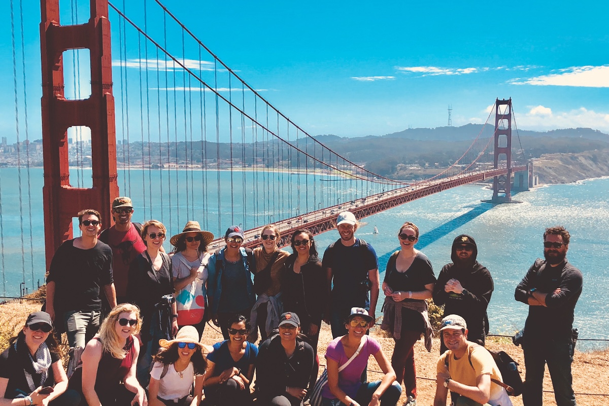 A group of people posing and smiling in front of the Golden Gate Bridge, with MarinCounty and San Francisco Bay visible in the background under a clear blue sky. The casually dressed group appears to be enjoying a bright, sunny day on one of their MarinTours.