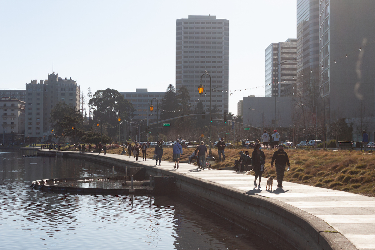 People walk along a waterfront path lined with trees and grass, some walking dogs. Tall buildings and streetlights frame the scene, while the sun shines brightly. The pathway curves along the edge of a body of water, inviting you to visit East Bay's scenic beauty.