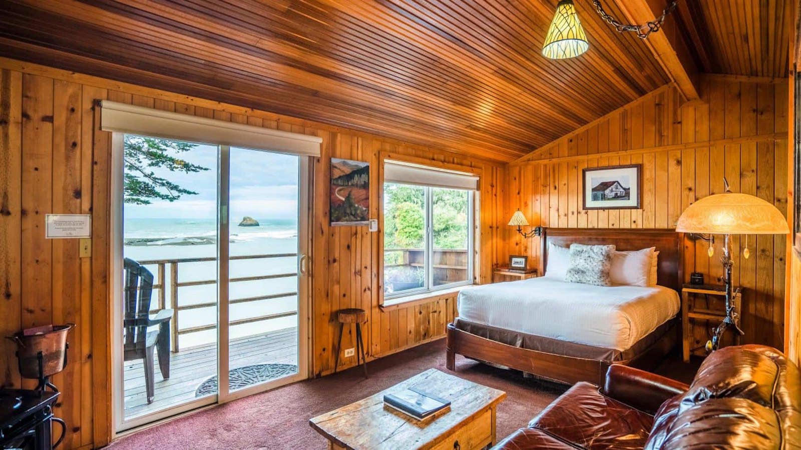 Cozy wooden cabin bedroom with a large window and sliding glass door opening to a balcony with an ocean view. A bed is placed against the wooden wall with a soft white duvet. A leather sofa and a wooden coffee table complete the warm, rustic decor, perfect for guests of luxury hotels on the Sonoma Coast.