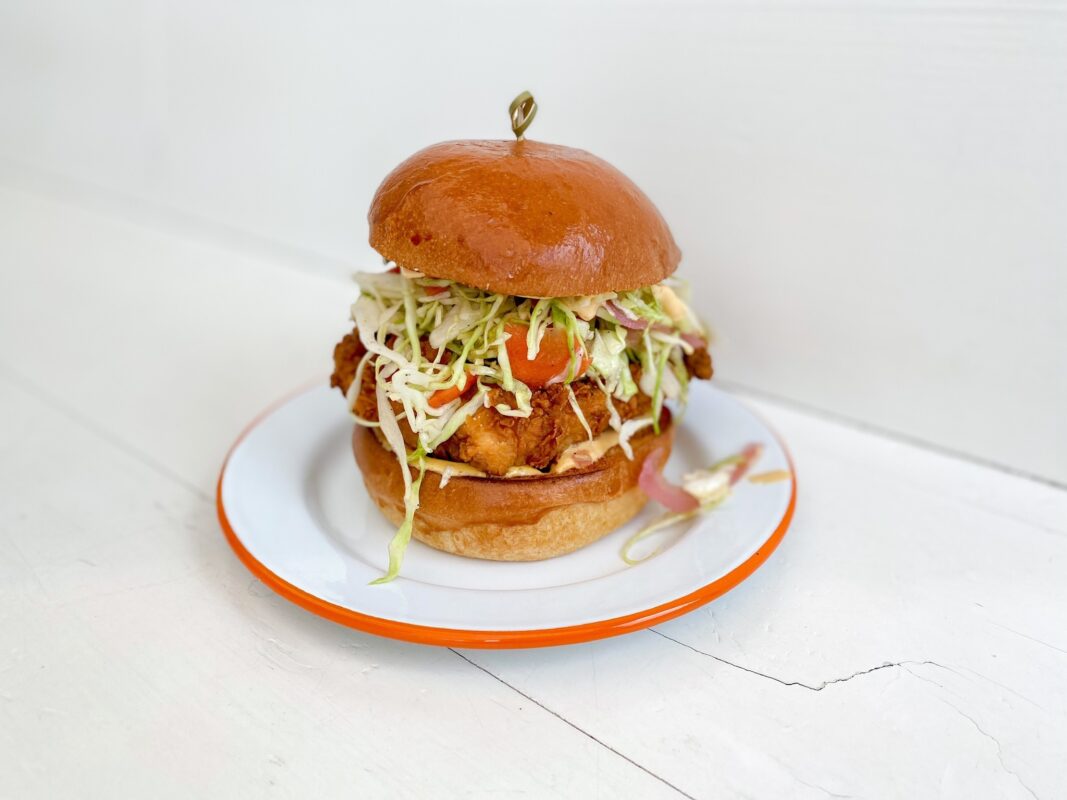 A fried chicken sandwich is presented on a white plate with an orange rim. The sandwich includes a fluffy bun, crispy fried chicken, shredded lettuce, sliced pickles, and a touch of sauce, held together with a toothpick. Enjoy one of the Best Burgers in Sonoma on a light wooden surface background.