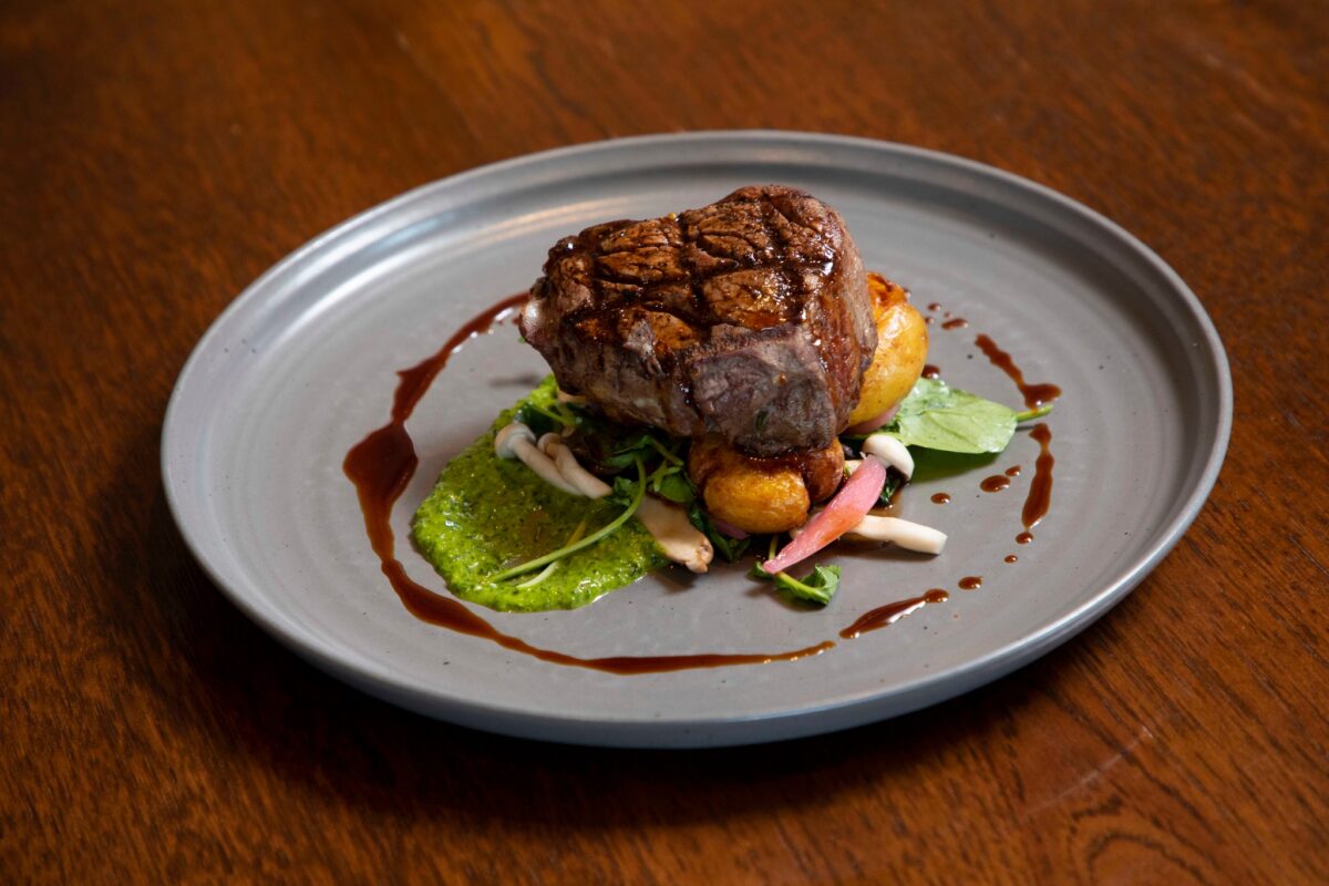 A gourmet dish featuring a perfectly-cooked steak resting on a bed of greens and roasted vegetables, garnished with a vibrant green sauce and drizzled with a rich, dark reduction. This elegant presentation is the Best Dinner on the Sonoma Coast, served on a simple gray plate placed on a wooden table.