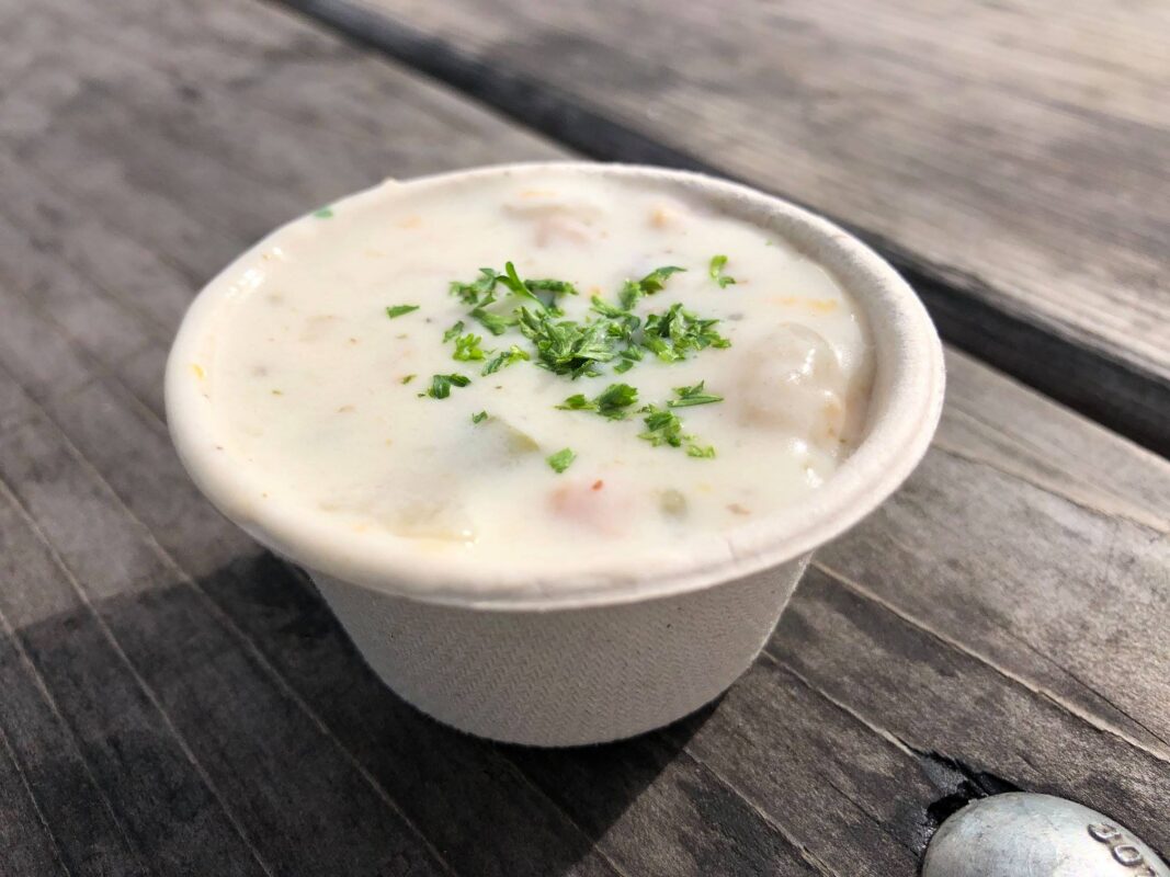 A small white paper cup filled with creamy soup, garnished with chopped parsley, sits on a wooden surface. The soup appears to contain small pieces of vegetables or seafood. A metal bolt is visible on the right edge of the image, capturing a moment reminiscent of the best lunch in Sonoma.