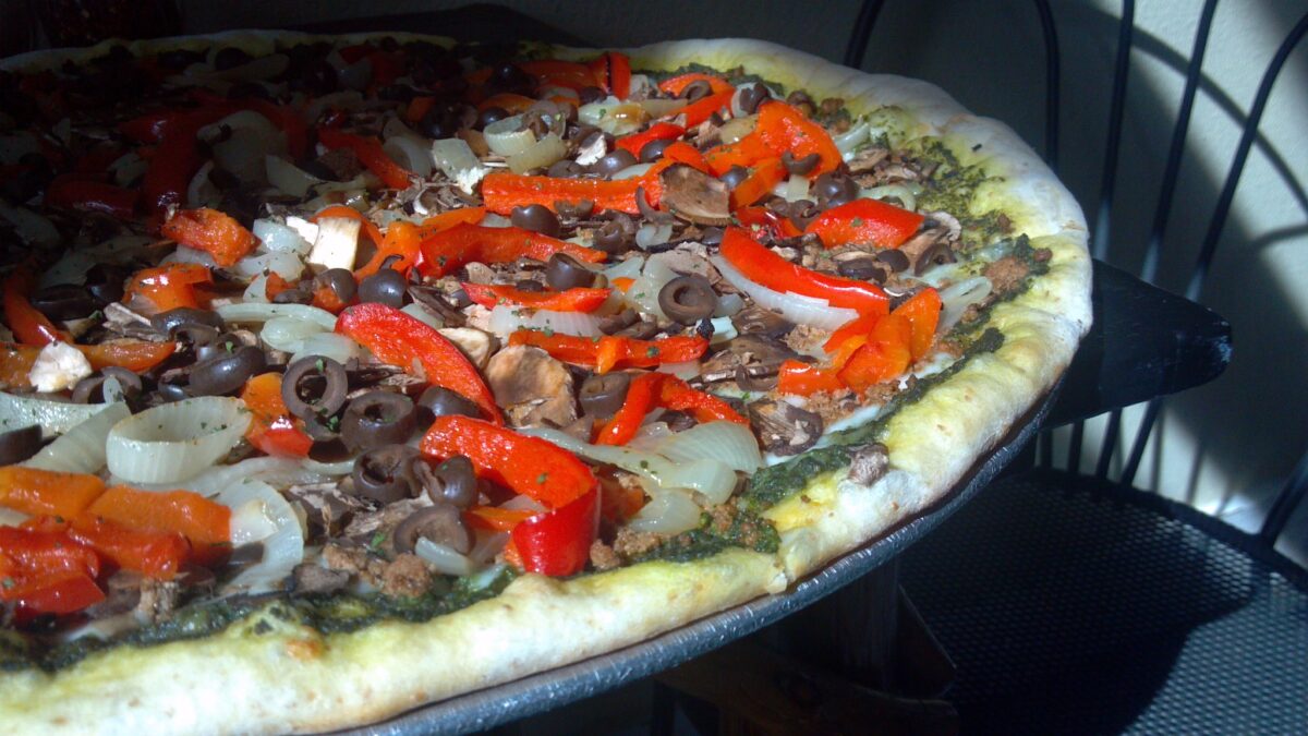 A close-up of the Best Pizza in Mendocino topped with red bell peppers, black olives, mushrooms, and onions. The pizza has a thick, lightly browned crust with a green pesto sauce base and is set on a wooden pizza stand in a well-lit room.