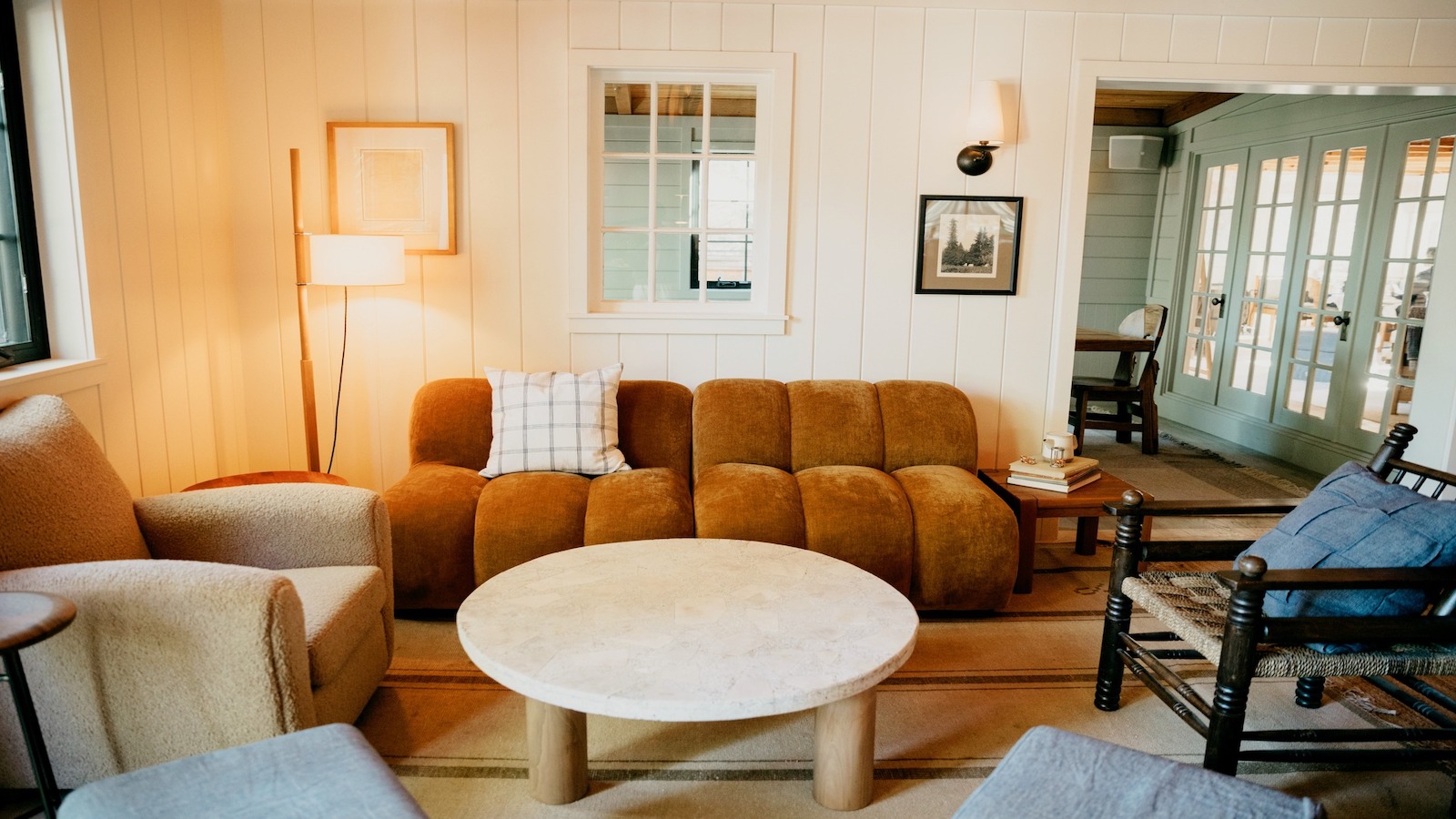 A cozy living room with a brown plush sofa, beige armchair, round coffee table, and mismatched chairs. The space is brightly lit with a floor lamp and wall sconce. Artwork and a window adorn the back wall, while glass-paneled doors lead to another room in this luxury hotel.