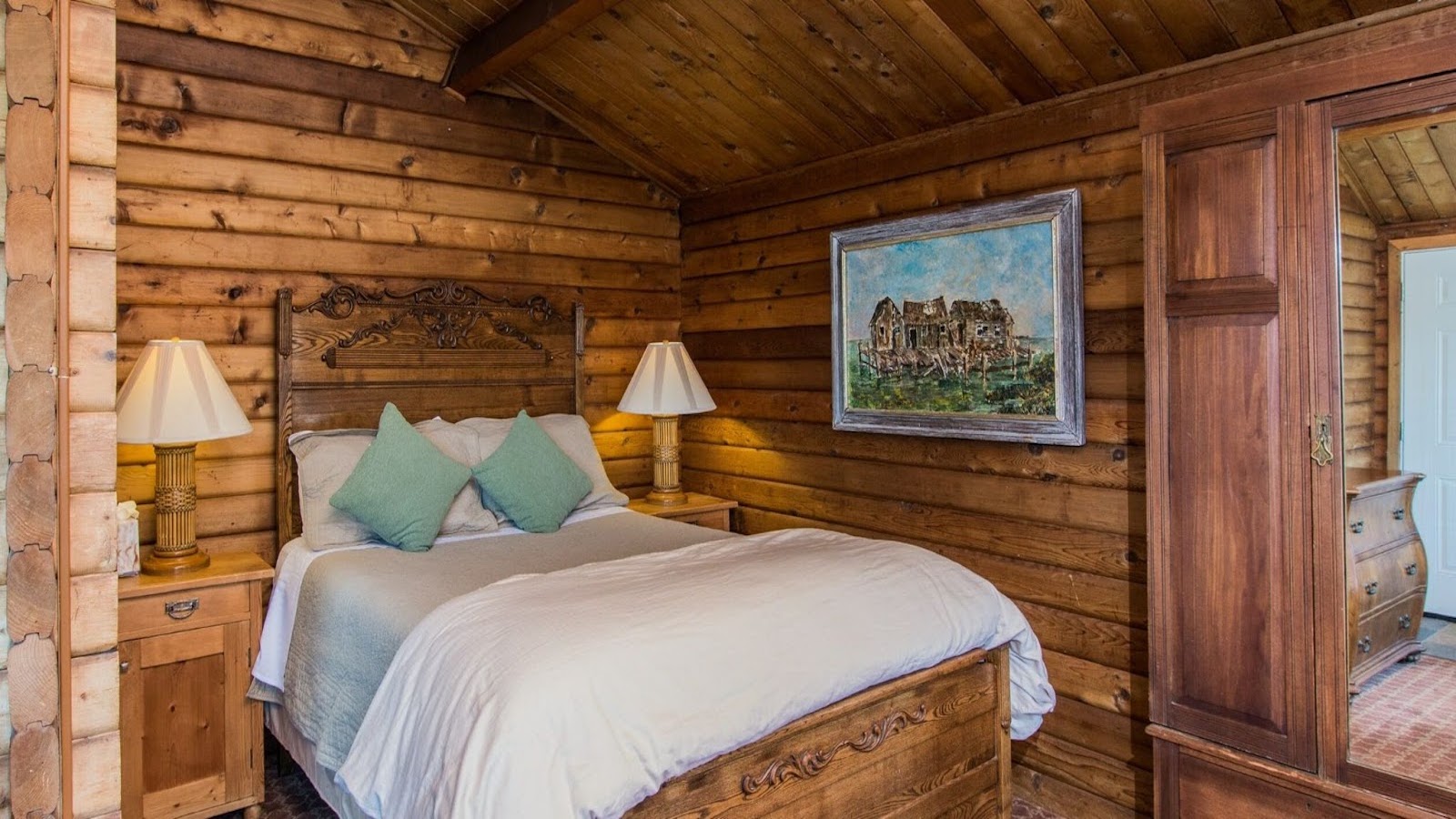 A cozy cabin bedroom with wooden walls and a pitched ceiling, reminiscent of luxury hotel suites at the best group getaways hotels Sonoma Coast. It features a wooden bed with white bedding and two green pillows. Two bedside tables with lamps flank the bed, and a large painting of a castle hangs on the wall. An armoire is placed to the right.