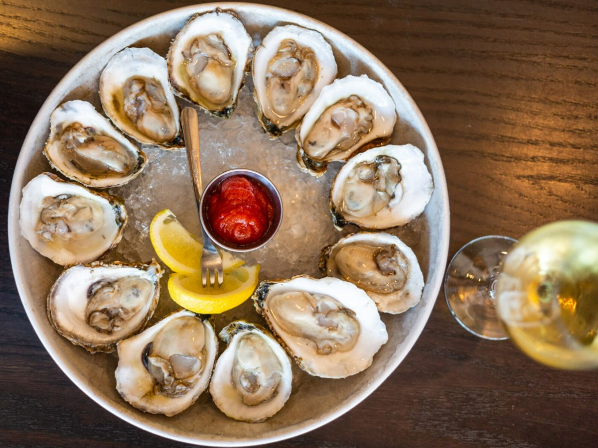 A round plate of oysters on the half shell is arranged in a circle on crushed ice. A small metal dish of red cocktail sauce with a spoon is in the center. Lemon wedges are placed at the front of the plate. A glass of white wine, perfect for enjoying the best lunch in Sonoma, is partially visible on the right.