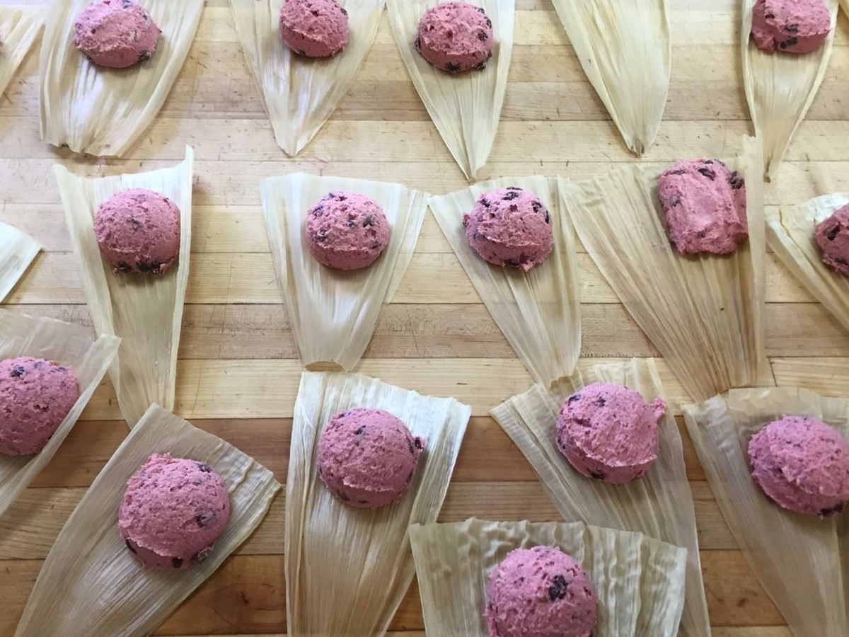 Corn husks laid out on a wooden surface, each topped with a portion of pink masa dough containing visible dark bits, likely to be used for making tamales—the best lunch in Mendocino. The dough portions are evenly spaced on the husks, ready for wrapping.