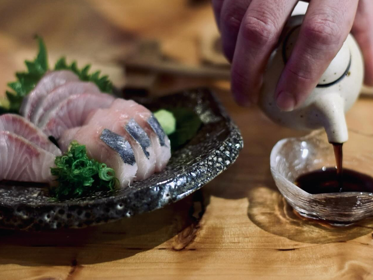 A close-up of a plate of sashimi garnished with greens on the left side of the image. On the right, a hand is pouring soy sauce from a small ceramic pitcher into a glass bowl. The scene, capturing the Best Sushi on the Sonoma Coast, is set on a wooden table.