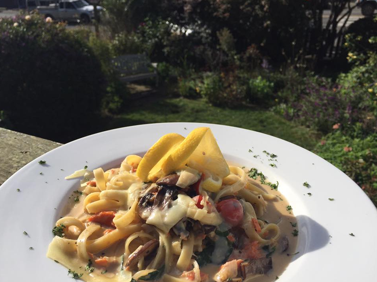 A plate of creamy pasta topped with shredded cheese and yellow squash slices is placed outdoors. The pasta is garnished with herbs and includes various ingredients like cherry tomatoes and greens. A garden and pathway are visible in the background, making it the best lunch in Mendocino.