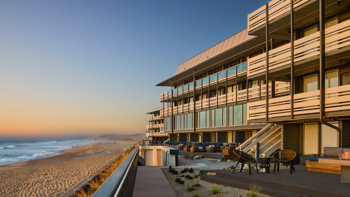 Modern beachfront building with balconies and large windows overlooking a sandy beach at sunset. The boardwalk of Monterey Beach Hotel features outdoor seating areas and planters, and the ocean's waves gently lap against the shore. Distant figures are seen walking along the beach.