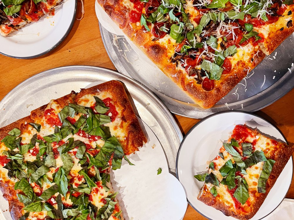 A table boasts the Best Pizza on the Sonoma Coast, featuring square-cut pizzas topped with fresh basil, melted cheese, and sliced pepperoni. The pizzas rest on silver trays, with some slices served on white plates in a casual dining setting.