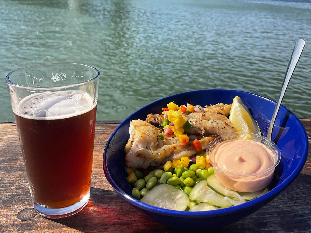 A glass of amber beer and a blue bowl filled with grilled fish, a lemon wedge, edamame, cucumber slices, and a small cup of dipping sauce are placed on a wooden surface with a body of water in the background. This delightful setup truly embodies the Best Lunch in Mendocino.