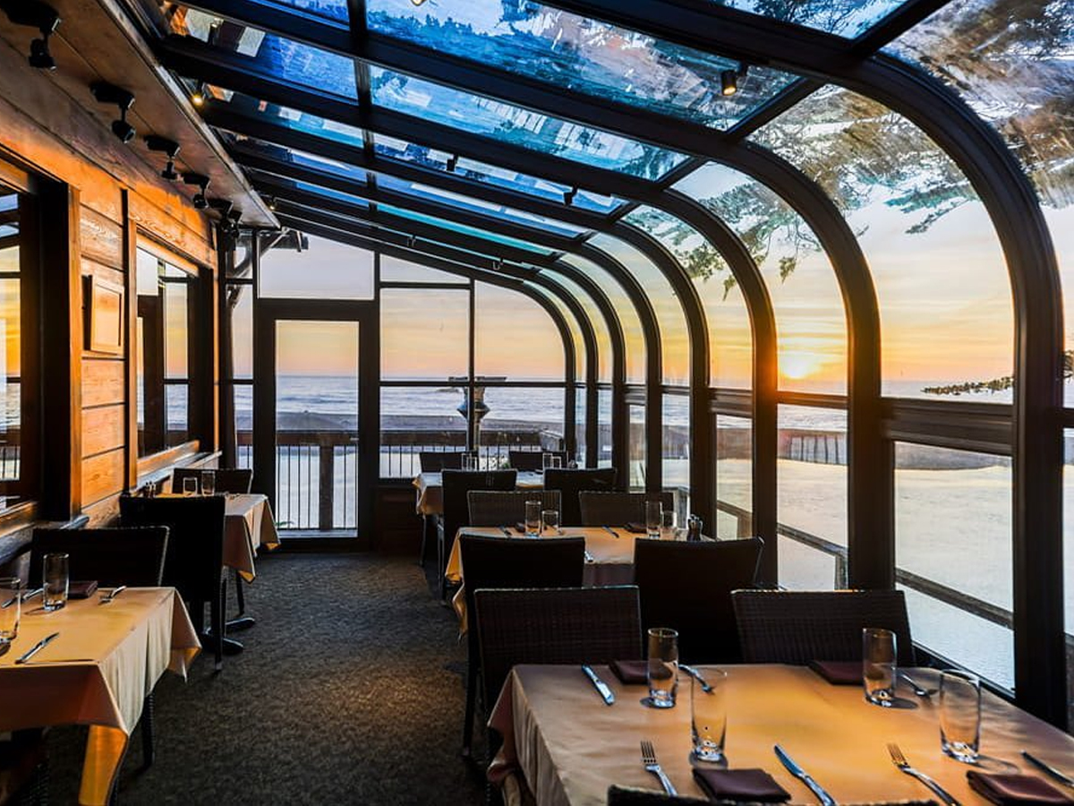 A cozy, glass-enclosed restaurant interior with neatly set tables and chairs is shown. The room offers a scenic view of the ocean at sunset through large windows, creating a warm, inviting atmosphere—perfect for the best dinner on the Sonoma Coast.