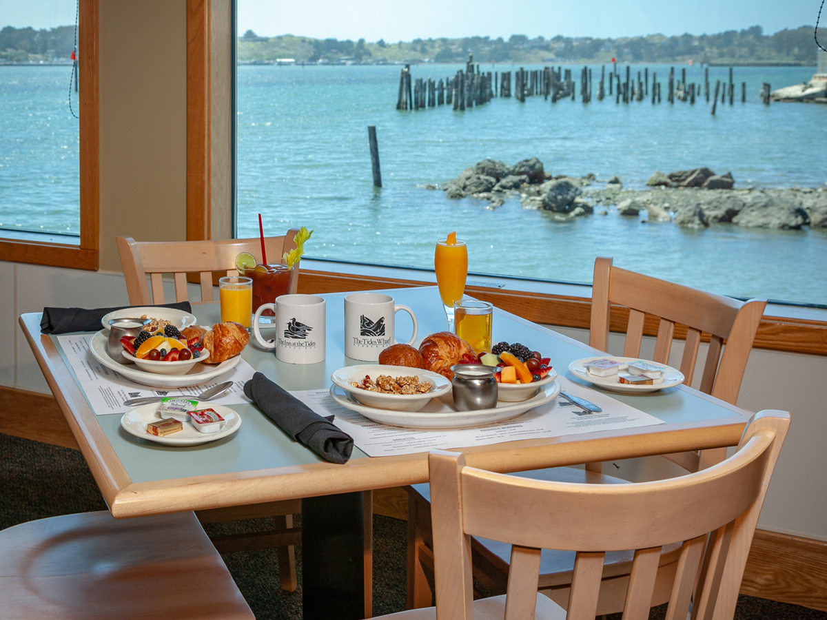 A dining table next to a window overlooking a scenic waterfront view. The table is set with various breakfast dishes, including pastries, fruit, cereal, and beverages like coffee and orange juice. Two wooden chairs are positioned around the table, reminiscent of the best dinner on the Sonoma Coast.