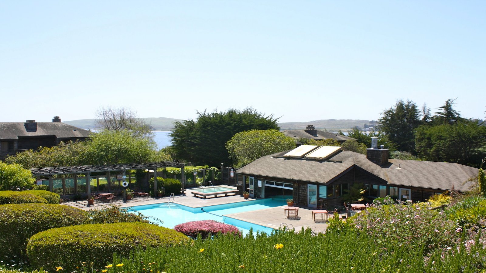 A scenic view of a luxurious house with a large swimming pool in the backyard, surrounded by well-manicured gardens. There's a pergola on one side, and the backdrop includes lush greenery, a distant body of water, and a clear blue sky, reminiscent of the best spa and wellness hotels Sonoma Coast offers.