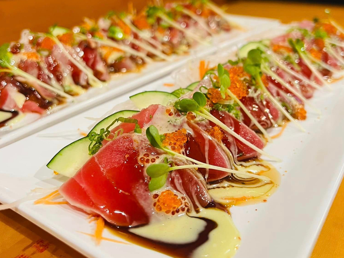 Close-up of a beautifully presented plate of sashimi. The dish, hailed as the Best Sushi on the Sonoma Coast, features fresh slices of raw fish, garnished with microgreens, cucumber slices, and fish roe, drizzled with a combination of creamy sauce and soy-based dressing, all arranged on a white rectangular plate.