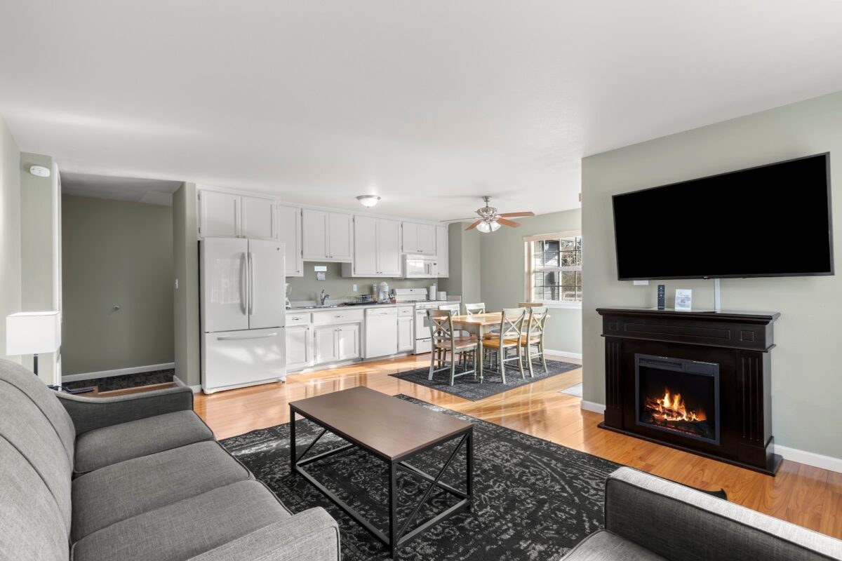 A modern living space with an open floor plan. The living area has a gray sofa, coffee table, dark rug, and a fireplace with a TV above it. Adjacent is the kitchen featuring white cabinets and appliances, a dining table with chairs, and light hardwood floors—ideal for those seeking luxury hotel vibes at home.