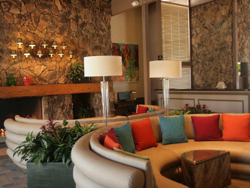 A hotel lobby with a cozy seating area featuring a semi-circle of beige couches adorned with colorful throw pillows in red, blue, and green. Two tall lamps and decorative plants sit on the table. The background features a stone wall with modern lighting, exemplifying the charm of the best value hotels Marin offers.