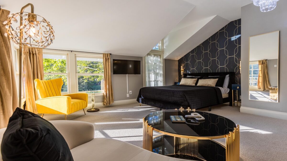 A modern bedroom in a luxury hotel with large windows features a double bed adorned with black bedding, a yellow lounge chair, and a sleek, round black coffee table with gold accents. The room also includes a black and gold accent wall, a flat-screen TV, and light tan curtains.