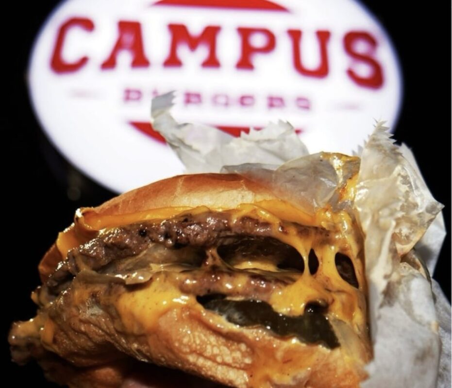 A close-up of a juicy cheeseburger with melted cheese and a beef patty, partially wrapped in white paper. In the background, there is a blurred sign that reads "Campus" in red letters on a white circular background, showcasing one of the top spots for where to eat in the Bay Area.