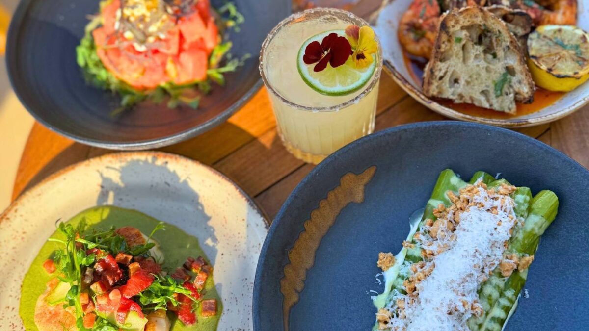 A table set with a variety of gourmet dishes and a cocktail promises the best dinner in Monterey. Plates include a salad with greens, a seafood dish topped with herbs, bread with grilled lemon, and asparagus with grated cheese and nuts. A margarita garnished with lime and a flower sits beautifully in the center.