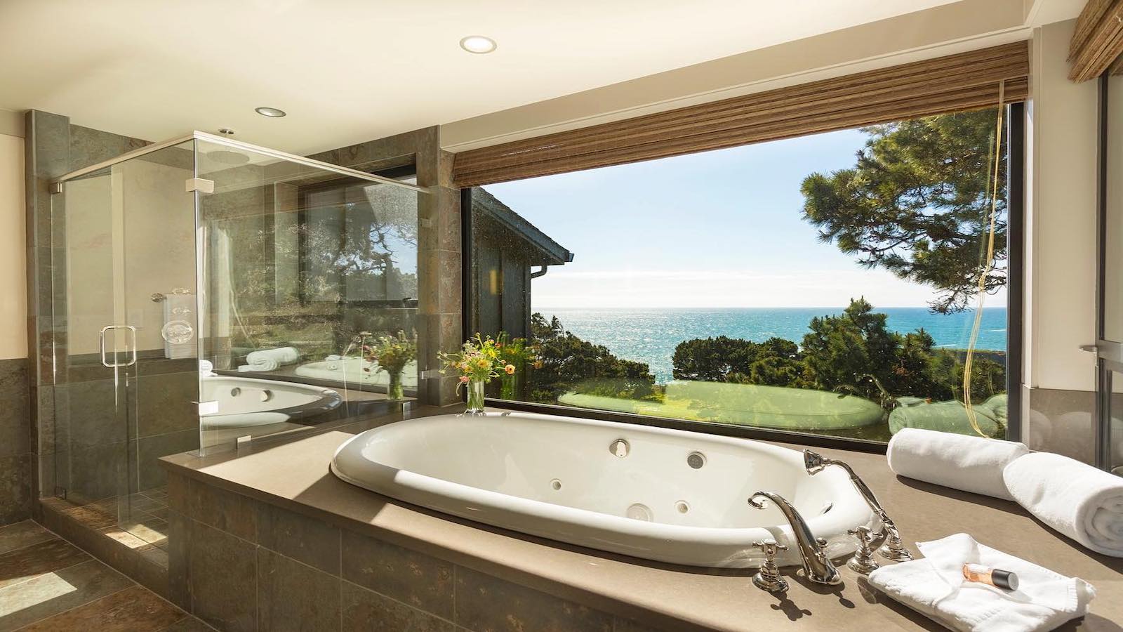 A luxurious bathroom in one of the best spa and wellness hotels Mendocino has to offer, featuring a large white bathtub set next to a window offering a stunning ocean view. The bathtub is surrounded by a stone-tiled platform, equipped with silver faucets, and adorned with fresh flowers and towels.
