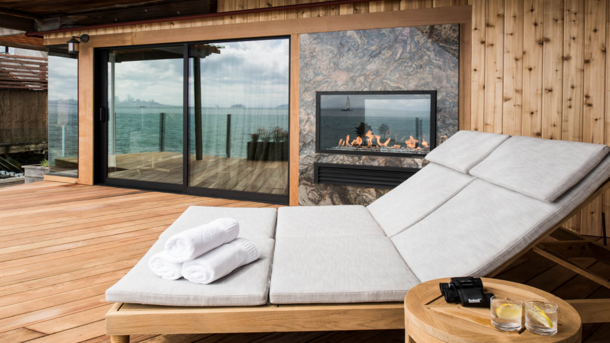 A cozy outdoor deck area with a cushioned chaise lounge, neatly folded towels, and a small wooden side table holding binoculars and two glasses of lemon water. The deck features a fireplace and sliding glass doors with a serene sea view in the background, reminiscent of the best wellness hotels in Marin.