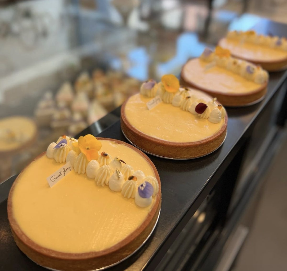 A row of four lemon tarts is displayed on a bakery counter. Each tart, topped with cream dollops and delicate edible flowers, epitomizes the culinary craftsmanship found in top "Where to Eat Bay Area" spots. In the background, more baked goods are partially visible out of focus.