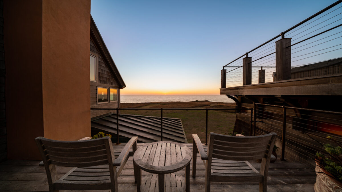 Two wooden chairs and a small table on a balcony overlook a serene ocean sunset. Framed by parts of two buildings, the sky transitions from blue to warm tones, reflecting on the calm sea, creating a tranquil and inviting atmosphere typical of the best group getaway hotels in Mendocino.