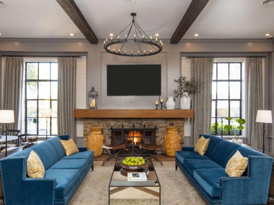 An elegant living room with two blue sofas facing each other, a wooden coffee table in between, and a fireplace with a mantel holding candles and plants. Large windows with curtains adorn the walls, and a round chandelier hangs from the ceiling above. For similar comfort, explore the best value hotels in Marin.