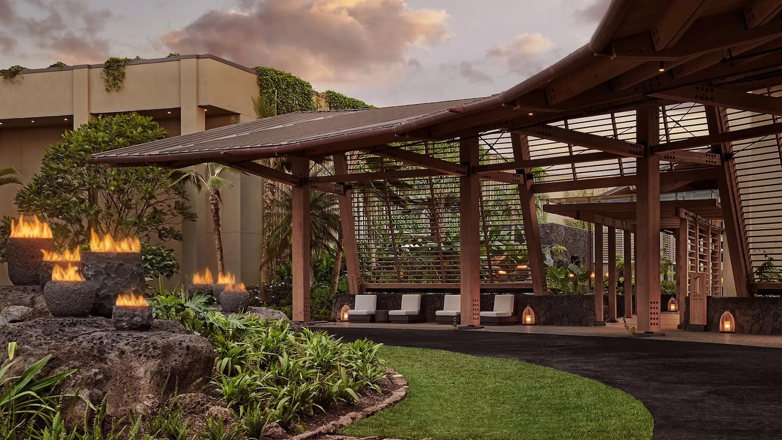 Outdoor area of a luxurious tropical resort in Hawaii featuring a covered walkway with wooden beams and roof, surrounded by lush greenery, decorative stone fire pits, and ambient lighting. Perfect for a romantic getaway, modern buildings partially covered with green ivy complete the backdrop.