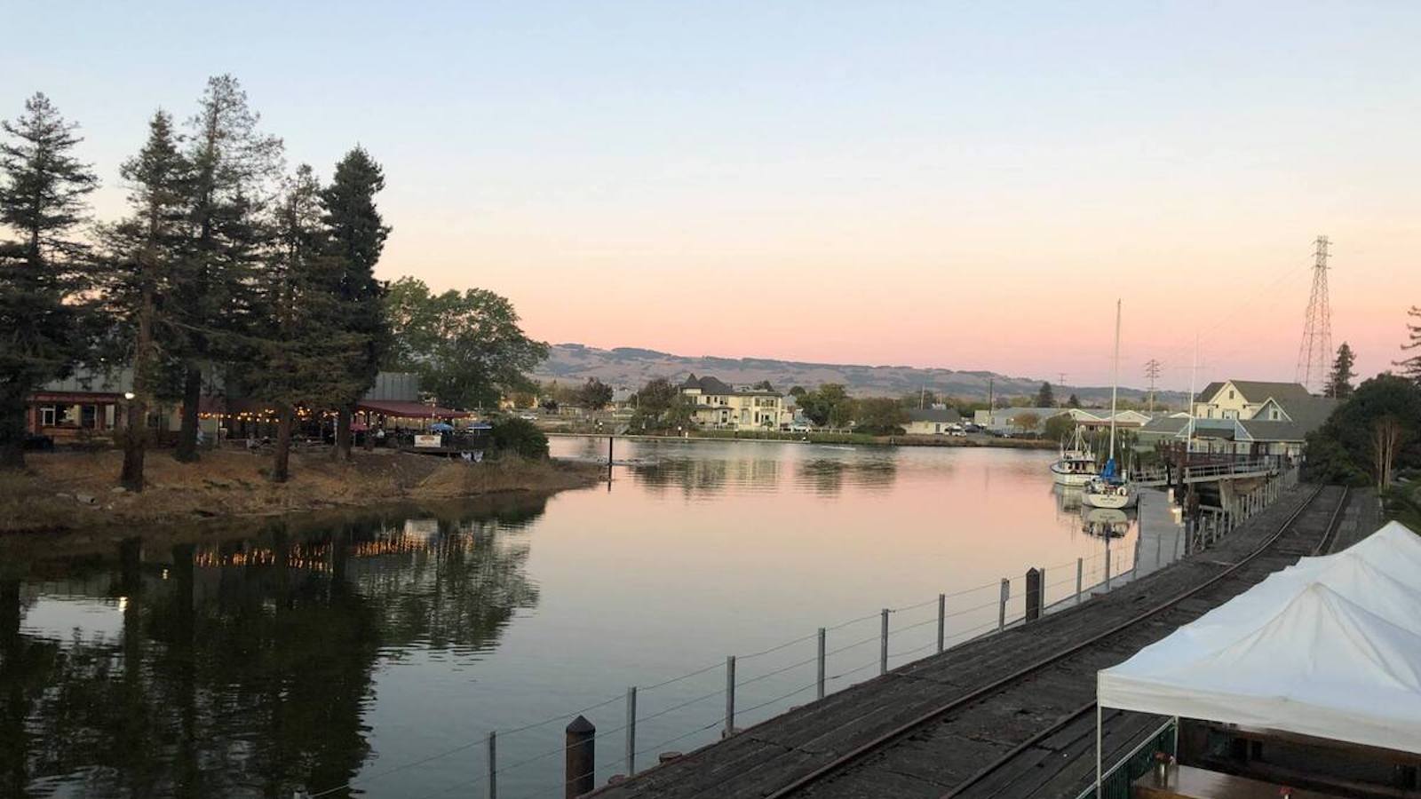 A scenic view of a calm, reflective river with boats docked on the right side. Trees line the riverbank on the left, and a few buildings, including houses and a dog-friendly tasting room with outdoor lights are visible. The sky is clear with a hint of pink from the setting sun.