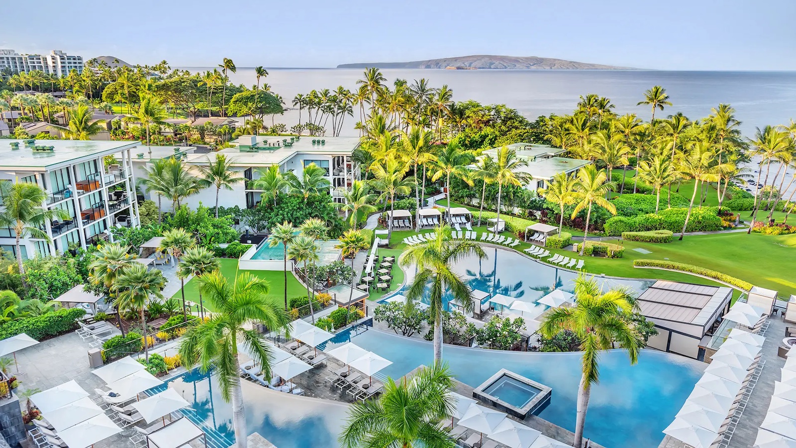 Aerial view of a luxurious tropical resort featuring multiple pools surrounded by palm trees, lounge chairs, and lush greenery. This ultimate luxury getaway in Hawaii boasts stunning resort buildings nestled among the foliage, with the tranquil ocean stretching towards a distant island in the background.