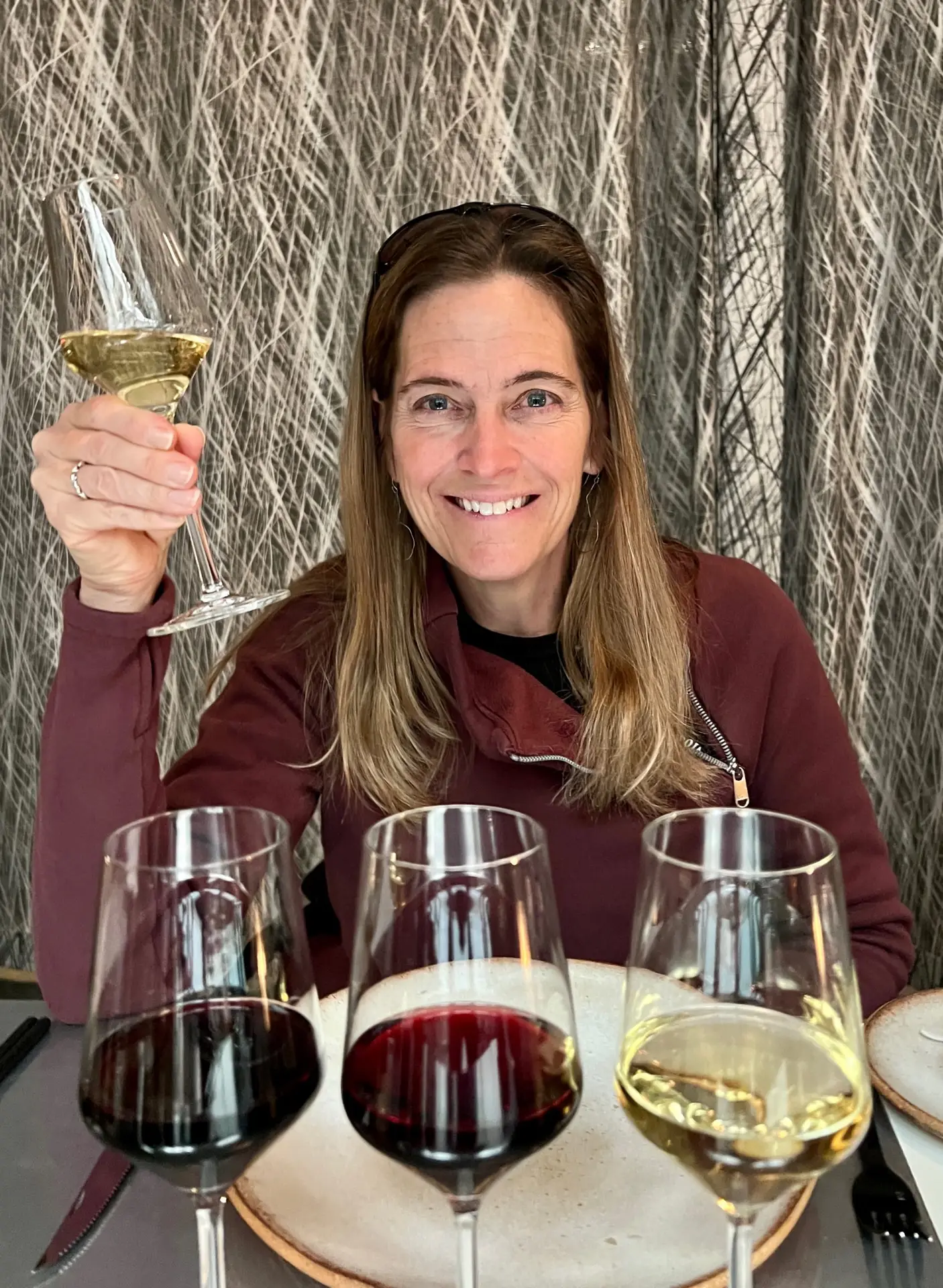 A woman with long brown hair smiles at the camera, holding up a glass of white wine in a toast. She is seated at a table with an empty plate in front of her, and there are three glasses of wine—two red and one white—on the table.