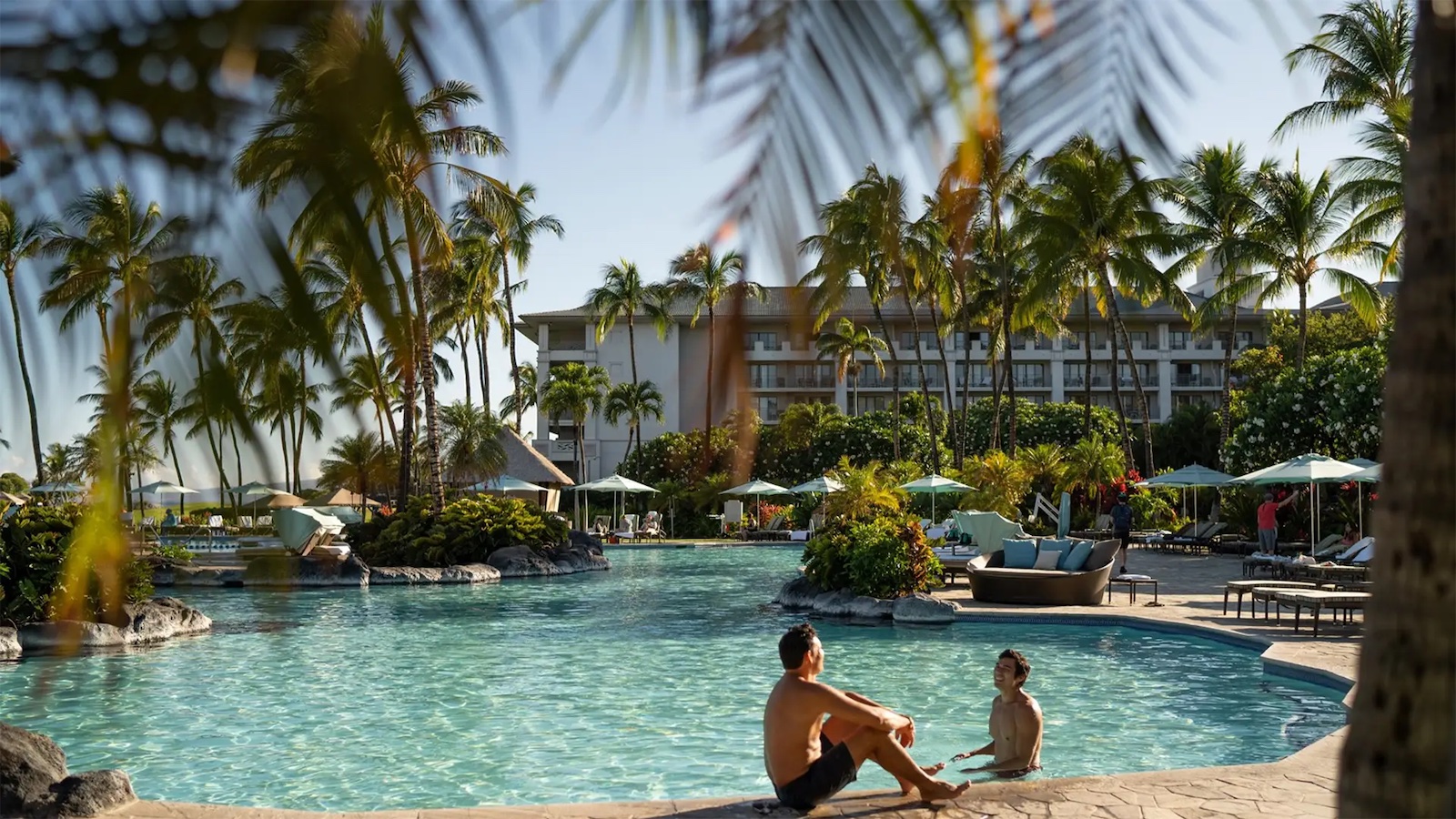 Two people sit by the edge of a large, tranquil pool surrounded by lush palm trees and greenery. Nearby, lounge chairs with umbrellas and a large building in the background can be seen under a clear blue sky. This could be one of those serene hotels with spas in Hawaii. A palm frond frames the top of the image.